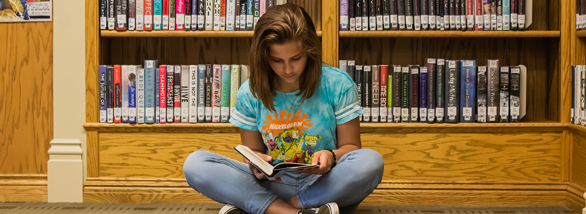 Teen reading a book while sitting on library floor