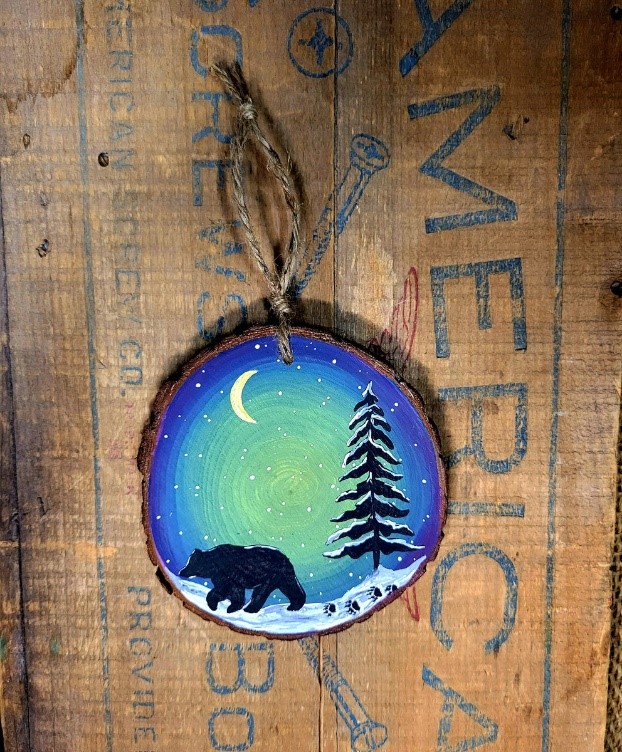 round wood slice painted with an outdoor scene, night sky, snow covered tree and a bear